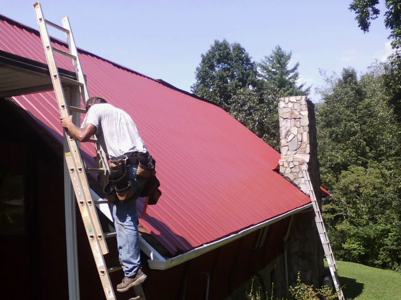 Roofer on a ladder beside a red metal roof and stone chimney — Soffit & Fascia Repair by Roofing Brothers in Rochelle Park